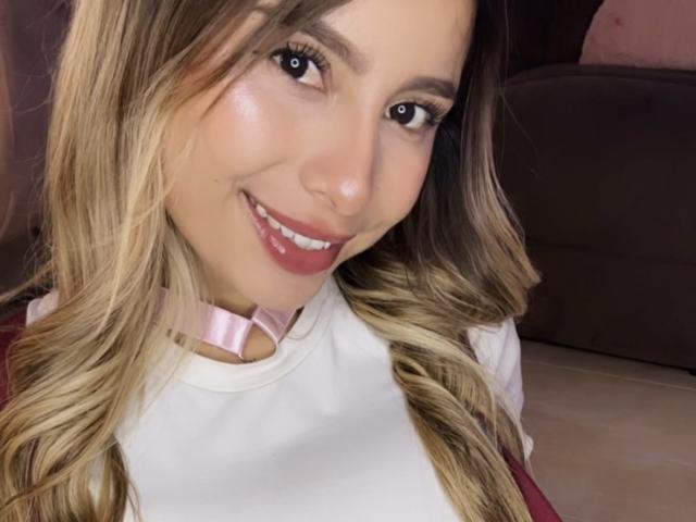 AmaraRicci tem cabelo loiro ondulado, olhos castanhos e um sorriso adorável com batom rosa. Ela está usando um colar rosa e uma blusa branca, que revelam seus seios levemente..