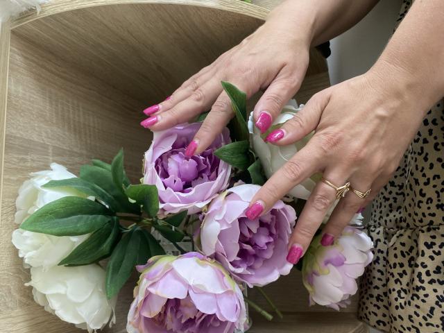Tilliana's hands with pink nails, showing rings, hold a bouquet of white and purple peonies with green leaves. Her leopard-print dress is partially visible.