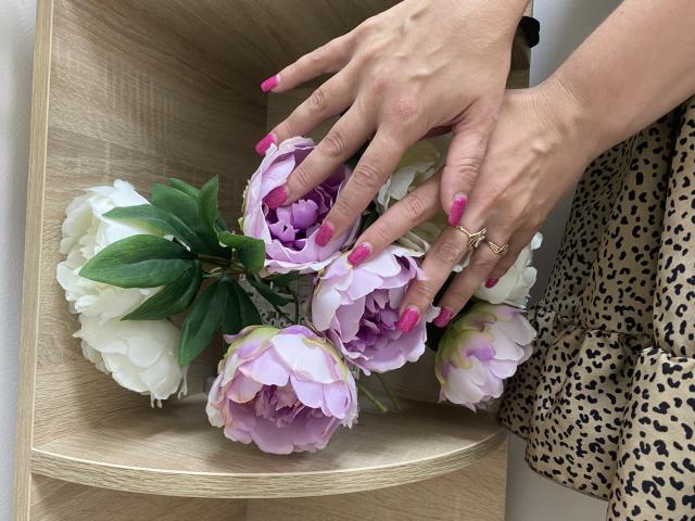 Tilliana's handen, met roze nagels en een gouden ring, houden roze en witte pioenbloemen vast op een houten plank. Ze draagt een jurk met luipaardprint..