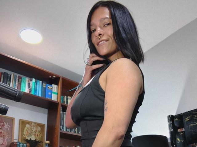 AmelieBlake with black hair, nose ring, and sleeveless black top, smiling, showing her tits, hand on chin, bookshelf and wall in background, looking cute and confident.