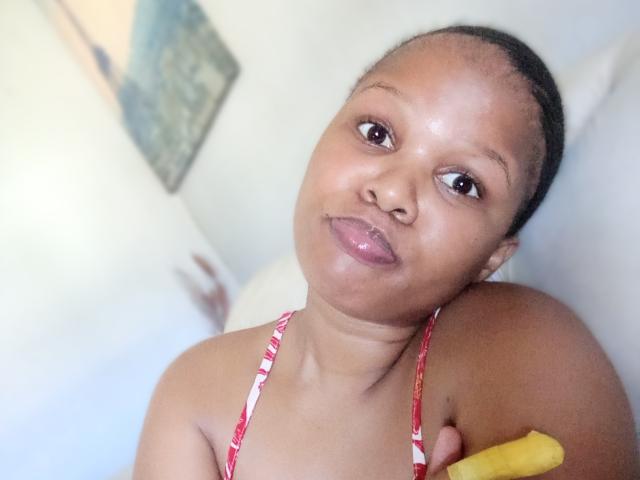Sammi Eshari, avec sa peau foncée, ses lèvres pulpeuses et un haut de bikini floral rouge, tenant une banane, regardant vers le haut avec un léger sourire, et avec des cheveux courts tressés..
