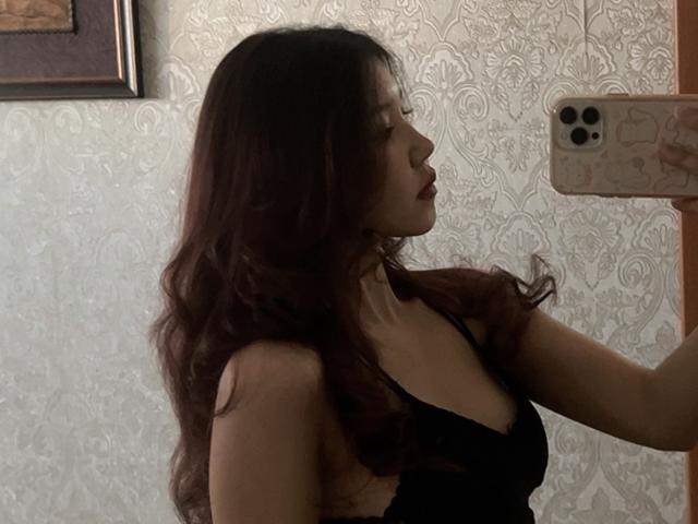 Elba Huppenbauer mit welligem, dunkelbraunem Haar, trägt eine schwarze Spitzenbluse und macht ein Selfie mit einem goldenen Handy. Dabei zeigt sie ihr Profil und ihre Brüste in einem schwach beleuchteten Raum..