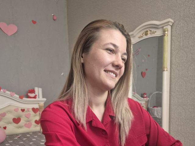 Anshante with blonde hair, smiling, wearing a red shirt, her ass slightly visible, standing in front of a mirror, pink heart decorations in the background.