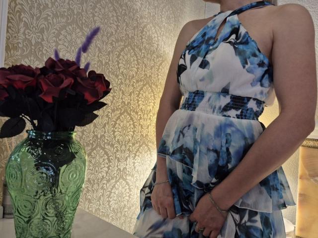 Anshante in a blue and white floral dress, showing her tits, stands next to a green vase with red roses and purple feathers.