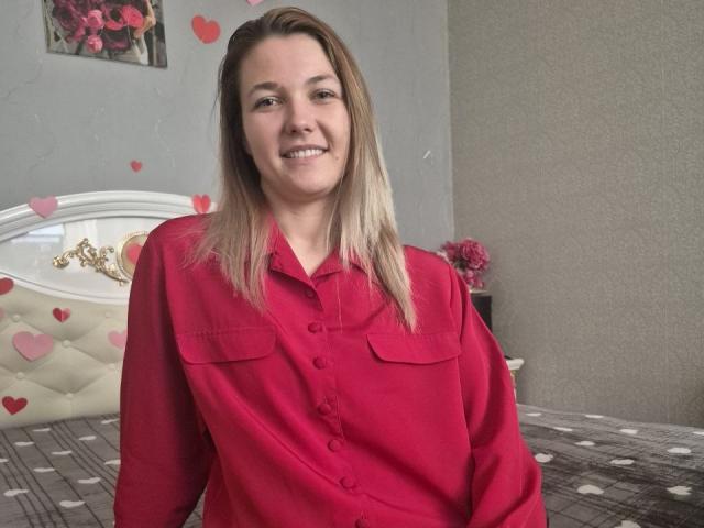 Anshante with blonde hair, smiling, wearing a red button-up shirt, standing in front of a bed with pink heart decals and a white tufted headboard.