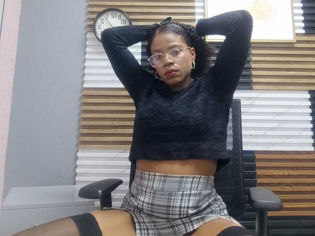 SarahiPusey with arms raised, wearing glasses, black sweater, plaid skirt, black thigh strap, and black leg garter, sitting on chair, looking serious. Clock in background.