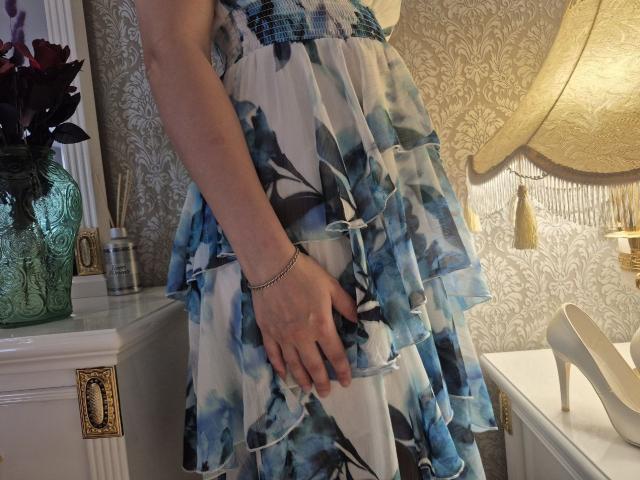 Anshante's hand rests on her blue and white floral dress with a ruffled skirt, wearing a silver bracelet, standing near white high heels and a green vase.