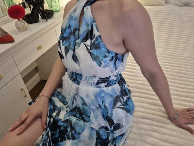 Anshante in a blue and white floral dress, sitting on a bed, showing her bare shoulders and side, with her hand on her thigh, and her ass visible.
