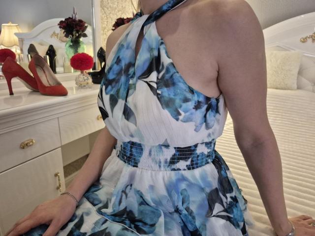 Anshante wearing a blue and white floral dress, halter top, showing her tits and arm, sitting on a bed, with red heels and a white dresser in the background.
