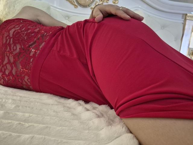 Anshante's red lace and fabric booty is on display, lying on a white bed with a fluffy texture. Her hand rests on her hip, showing a ring.