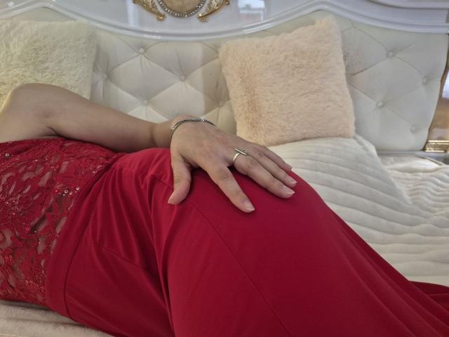 Anshante's hand with a ring rests on her red dress-covered ass, lying on a white tufted bed with beige and pink pillows. Her lace-covered thigh is visible.