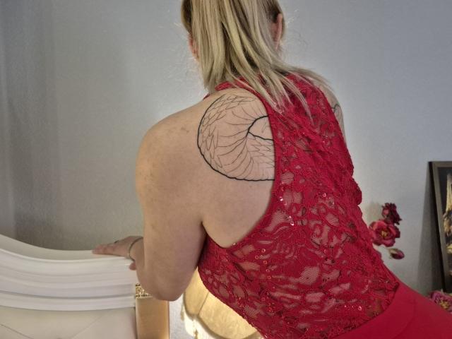 Anshante with blonde hair in a ponytail, wearing a red lace backless top, showing a black tree tattoo on her right shoulder, leaning on a white bed.