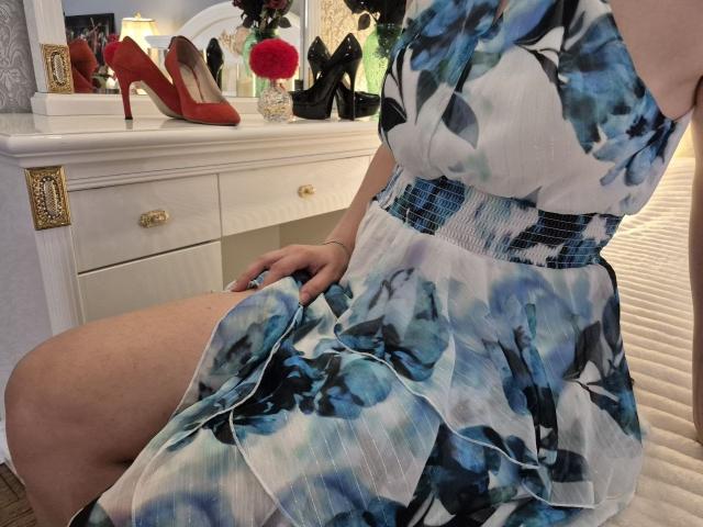 Anshante in a blue floral dress with a black and blue belt, sitting on a bed, showing her thighs and hand on her dress, red high heels on a white dresser.