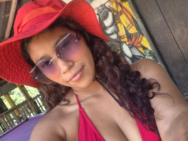 SeaHot, con i capelli castano ricci, un cappello da sole rosso, occhiali da sole rosa e un top da bikini rosa, mostra il seno, sorride e guarda la fotocamera..