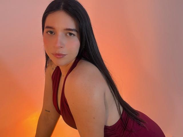 Valentina Riva, met lang zwart haar, draagt een rode halterjurk, leunt voorover, toont kleine borsten, heeft een subtiele tatoeage op haar linkerarm, zachte oranje belichting, licht open lippen, kijkt naar de camera..