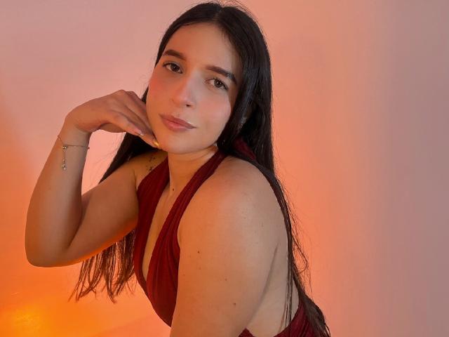 Valentina Riva, met lang zwart haar, draagt een rode halterjurk, heeft kleine borsten, een schattig gezicht en een subtiele glimlach, terwijl ze haar kin aanraakt, in een warm oranje licht..