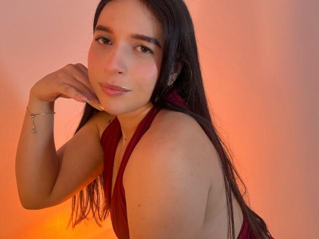 Valentina Riva, met lang donker haar, draagt een rode haltertop, leunt haar kin op haar hand, glimlacht zachtjes en draagt een armband met een klein hangertje, in een warm oranje licht..