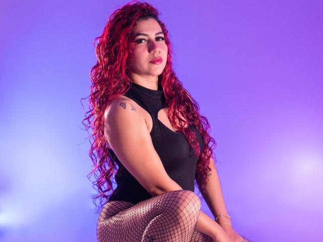 Antonela Riveiro met rood krullend haar, een zwart topje zonder mouwen, tatoeages op haar schouder, netbroek, zittend, paars-blauwe achtergrond, zelfverzekerde uitdrukking, roze lippenstift, sexy, volslank, volle borsten, subtiele make-up..