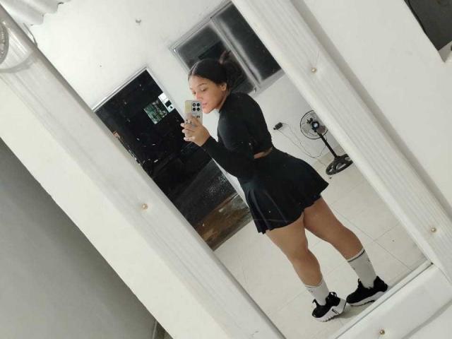 SheilaBrunette with black top, skirt, white socks, and black shoes, taking a mirror selfie, showing her ass and small tits, standing in a white room.