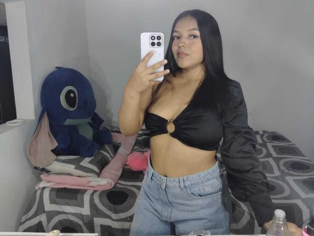 SheilaBrunette with long black hair, wearing a black velour bra with gold ring, showing her tits, in high-waisted jeans, holding a phone, next to a blue Stitch plushie.