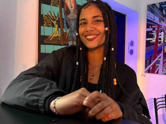 CinthiaWalker, smiling with brown skin and long braids, wearing a black jacket, has small tits, a nose ring, and a bracelet, sitting at a table.