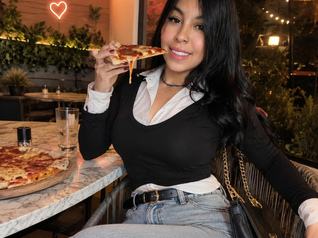 NenaBlack with black hair, medium tits, in a black top and blue jeans, eats pizza, holding a slice to her mouth, smiling, sitting at a marble table, night.