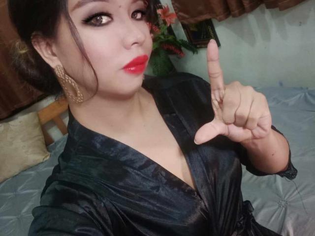 MistressAdriana, top rouge, haut en soie noire et boucles d'oreilles dorées, pointant du doigt, montrant son sourire éclatant et ses seins légèrement visibles dans un selfie pris dans une chambre..