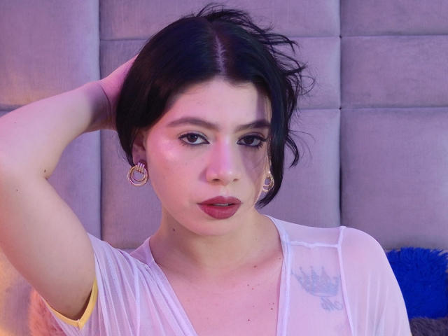 LindaMancini with short black hair, wearing a sheer white top, has her hand in her hair, showing off gold hoop earrings and red lipstick, with a subtle pink glow.