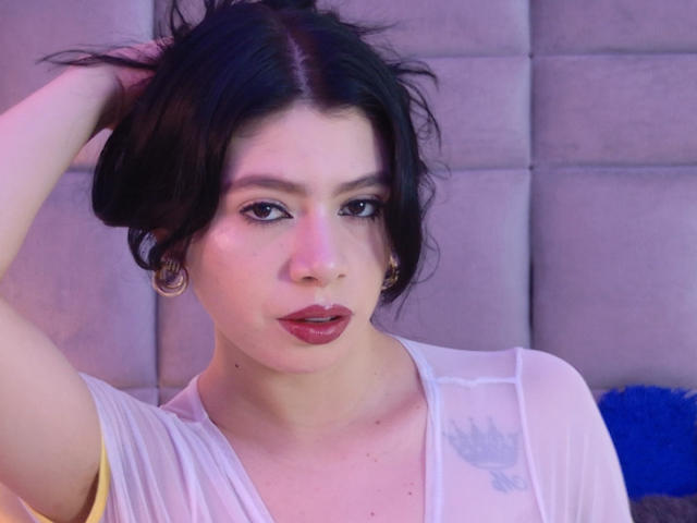 LindaMancini with dark hair, red lipstick, and a white sheer top, holding her hair up, showing off her small tits, and wearing gold hoop earrings.