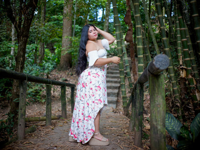 ElizaWiliamst è in piedi su un sentiero di legno, con una mano tra i capelli, indossa una maglietta bianca con una spalla scoperta e una gonna floreale, con sandali, mostrando i suoi seni e i suoi glutei in una foresta di bambù..