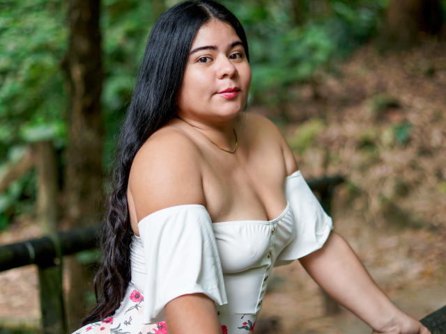 ElizaWiliamst, con lunghi capelli neri, una maglia bianca a spalle scoperte e un abito floreale, che mostra il seno, seduta in una foresta, sorride leggermente, indossando una collana d'oro..