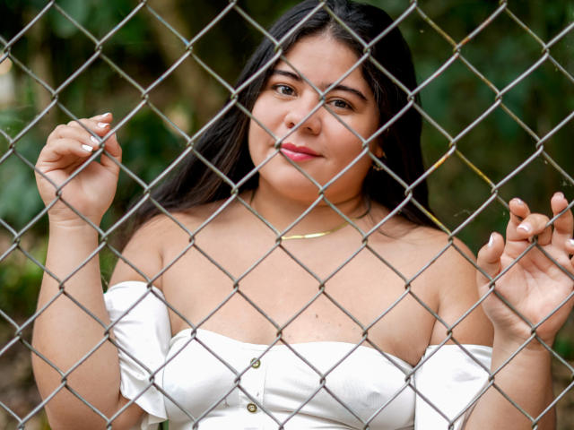 ElizaWiliamst with dark hair, white off-shoulder top, small gold necklace, holding chain link fence, showing her tits, smiling slightly, green background.