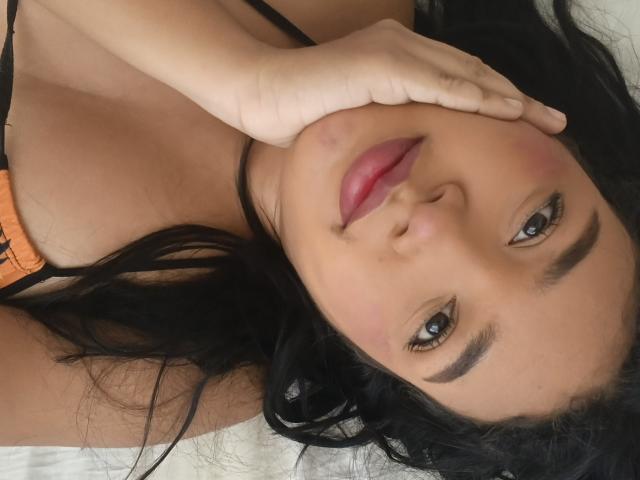 Katyi69 with dark hair, blue eyes, and pink lipstick, touching her cheek with her hand, wearing an orange top, showing her small tits, lying down.