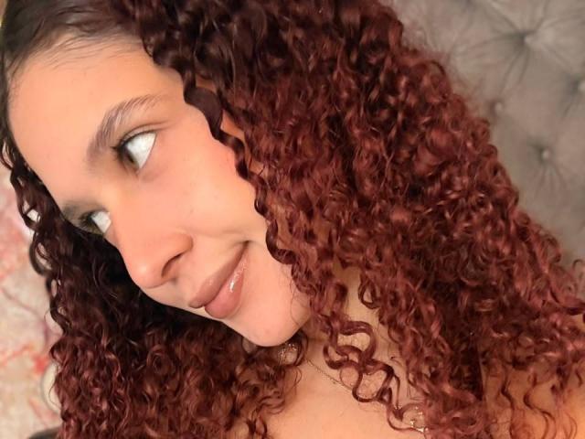 ValeryScot with curly red hair, smiling at the camera, looking to the side with green eyes, wearing a necklace, lying on a tufted grey surface.