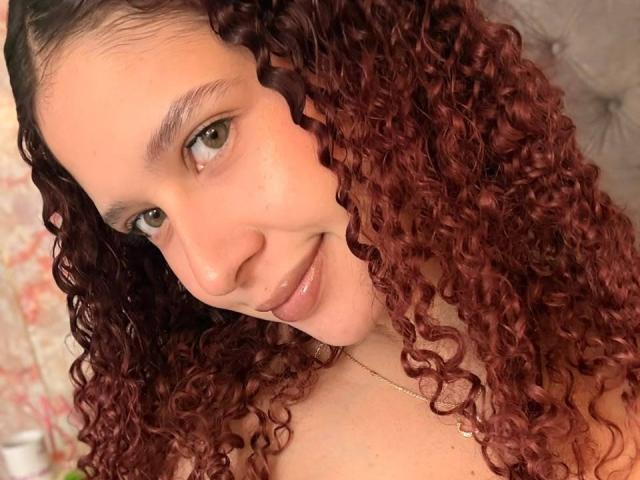 ValeryScot has curly red hair, light brown eyes, and a subtle smile. Her bare shoulders are visible, and she's wearing a thin gold necklace. Her skin is fair.