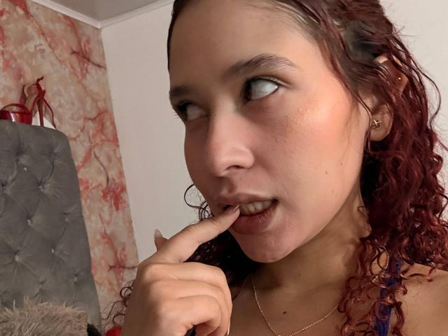 ValeryScot with curly red hair, fair skin, and a gold necklace, biting her finger with a slightly open mouth, looking to the side. Her tits are visible.