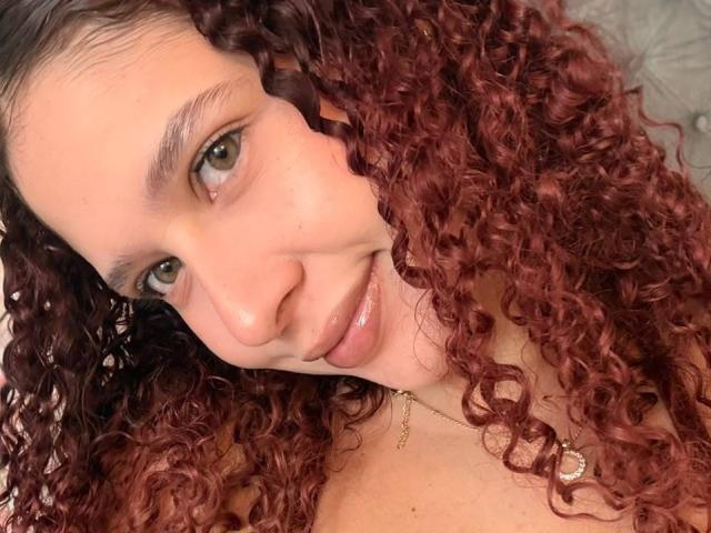 ValeryScot has curly red hair, green eyes, and a cute smile. Her nose is slightly upturned, and she's wearing a necklace with a small pendant.
