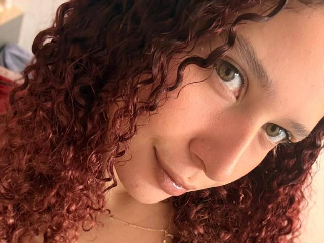 ValeryScot with curly red hair, light brown eyes, and a slight smile, wearing a gold necklace, looking directly at the camera. Her face is centered in the shot.
