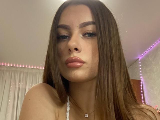 KimberlyZampieri has glossy pink lips, long brown hair, and blue eyes, wearing a white lace top with a small pendant, in a soft-lit room with pink LED lights.