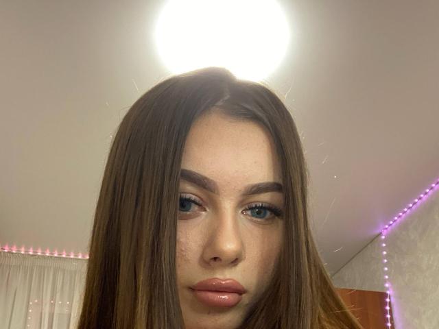 Het gezicht van Kimberly Zampieri, met lang bruin haar, blauwe ogen en glanzende roze lippen, onder een helder plafondlicht, met paarse LED-strips op de achtergrond..