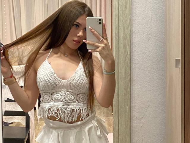 Kimberly Zampieri, met lang bruin haar, dat ze vasthoudt, draagt een witte gehaakte top, waarbij haar kleine borsten zichtbaar zijn, een witte rok, en ze maakt een spiegelzelfportret met een iPhone..