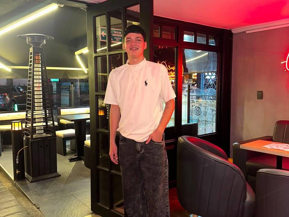 AlejoBussnekk está em um bar com pouca luz, vestindo uma camiseta branca com um logotipo preto, calças jeans escuras e um boné preto, sorrindo com as mãos nos bolsos..