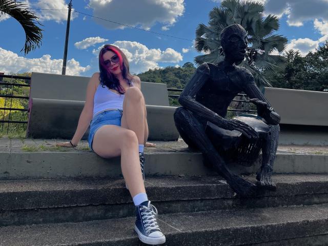 MaraCortes met roze en blauw haar, een wit tanktop, denim shorts en blauwe sneakers, zittend op betonnen treden naast een zwarte, metalen sculptuur van een zittende figuur..