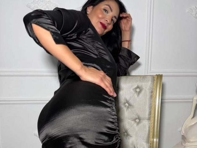 AlizonMoreti in a shiny black dress, leaning on a tufted chair, showing her ass, hand on hip, black hair, red lipstick, confident, sexy, looking at camera.