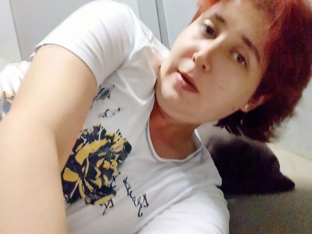 Lucy-2026-hot with short red hair, white t-shirt, and small tits, lying on a bed, taking a selfie with a slightly open mouth, looking at the camera.