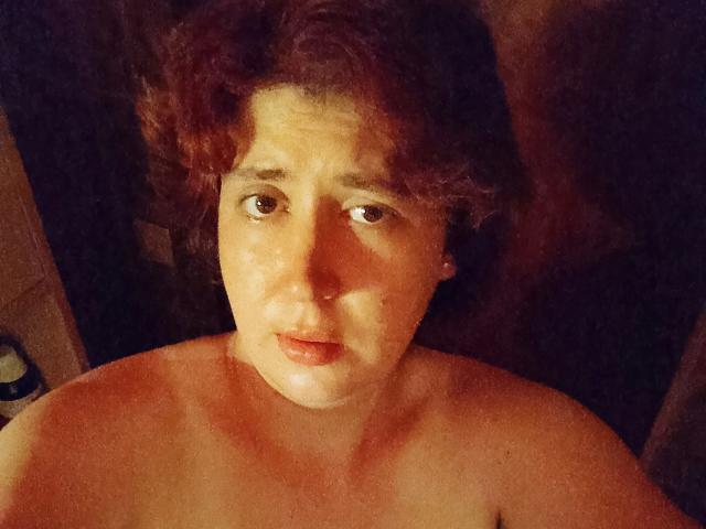 Lucy-2026-hot with shoulder-length curly brown hair, looking up with a slight smile, nude, brown eyes, light skin, small nose, and pearl earrings, in a dimly lit room.