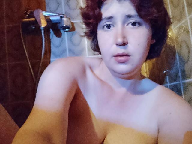Lucy-2026-hot tiene el pelo corto, rizado y rojo, piel clara, y está desnuda, con senos pequeños visibles, en un baño, sosteniendo un cabezal de ducha en un selfie..