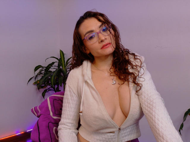 Drikka with curly brown hair, purple glasses, white zip-up jacket, deep cleavage, showing big tits, necklace, and a slightly bored expression. Potted plant and purple blanket in background.