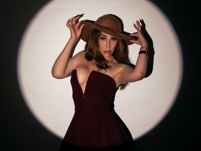 IssaMilfX with tan hat, hands on hat, dark maroon strapless dress, deep cleavage, brown eyes, long brown hair, black nail polish, white spotlight, black wristwatch.
