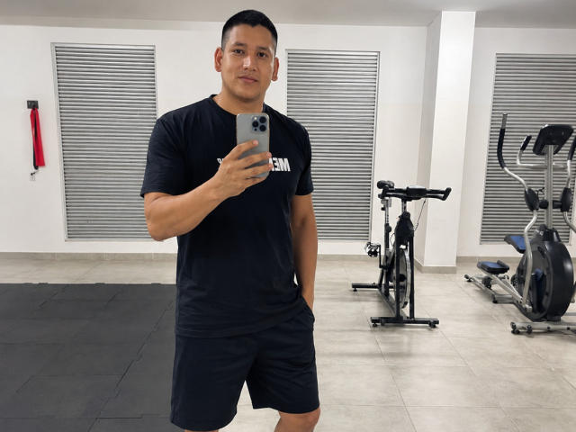 DanteVega è in una palestra, indossa una maglietta nera e pantaloncini, tiene in mano uno smartphone, ha una corporatura muscolosa, capelli neri corti e un sorriso sicuro..